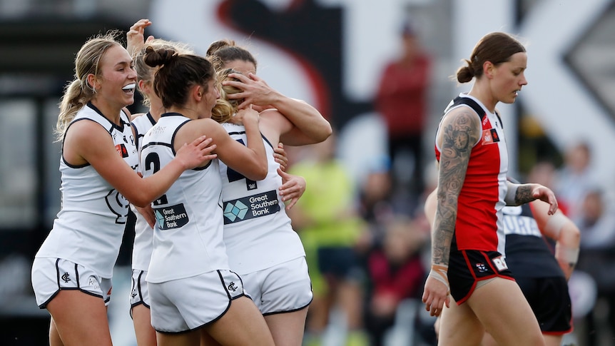 Four Carlton AFLW players embrace as they celebrate a goal while a St Kilda opponent walks near them.