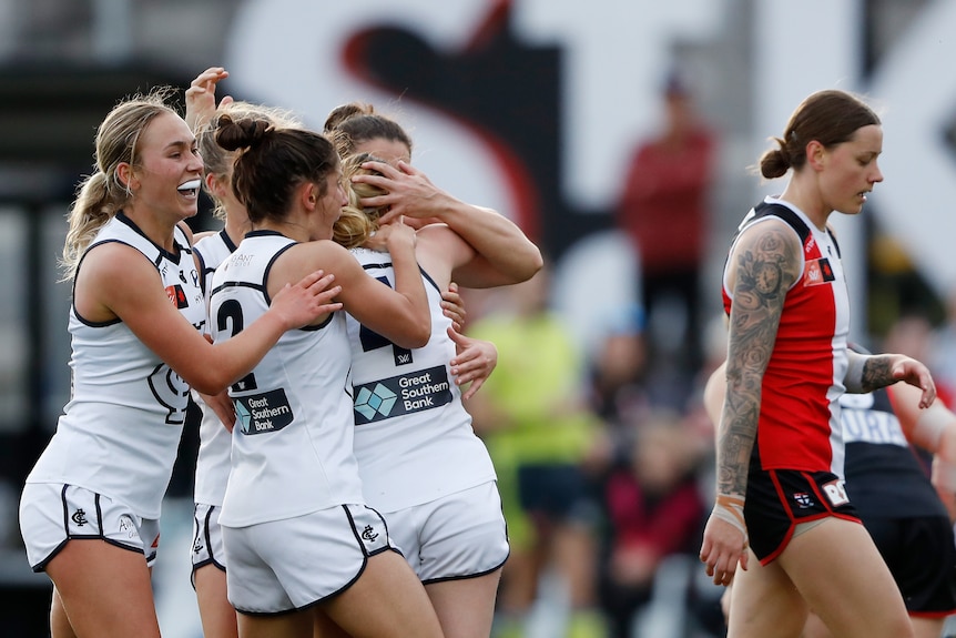 Four Carlton AFLW players embrace as they celebrate a goal while a St Kilda opponent walks near them.