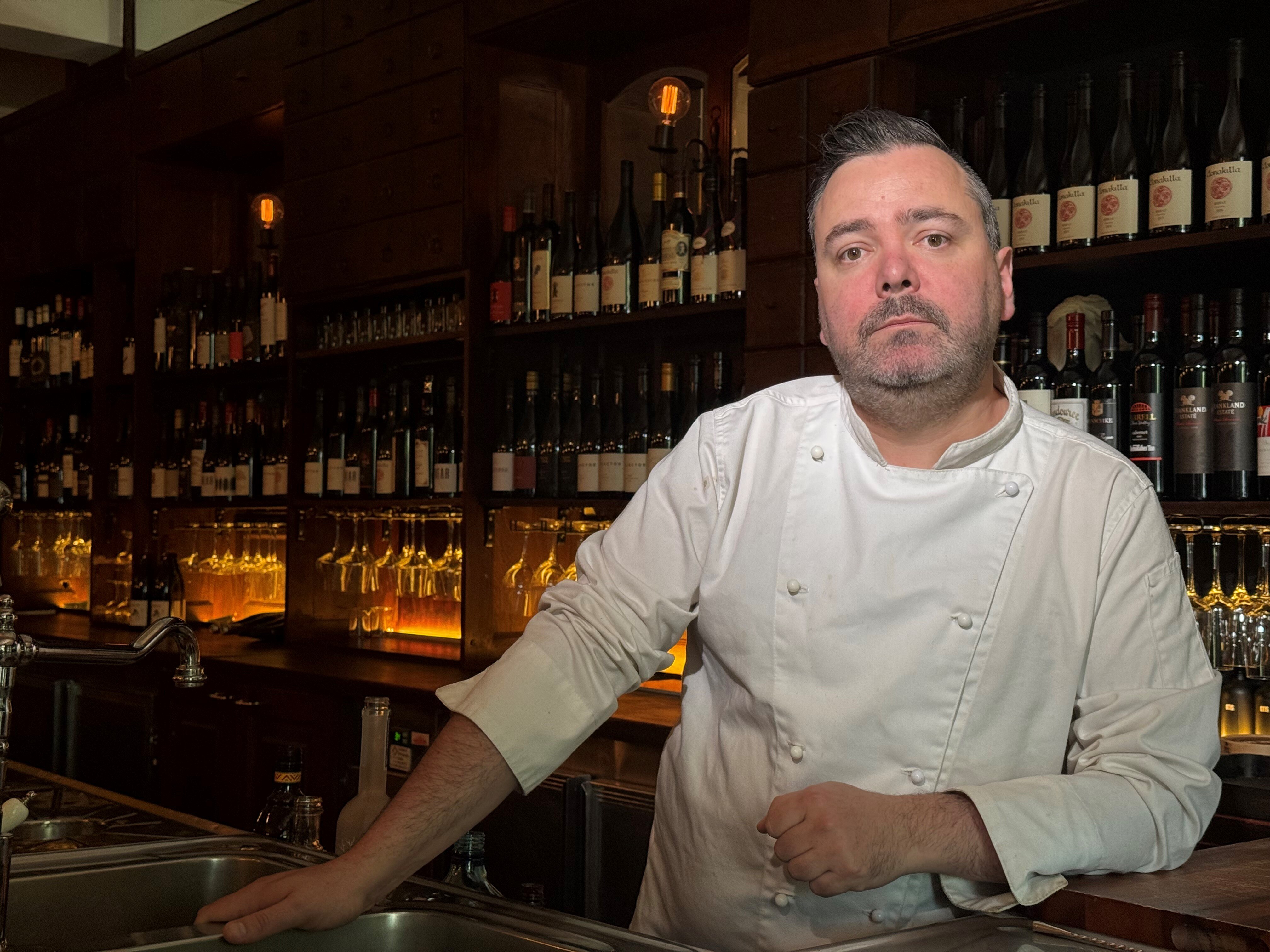 A man wearing a chef's outfit stands behind a bar in front of many bottles of wine.
