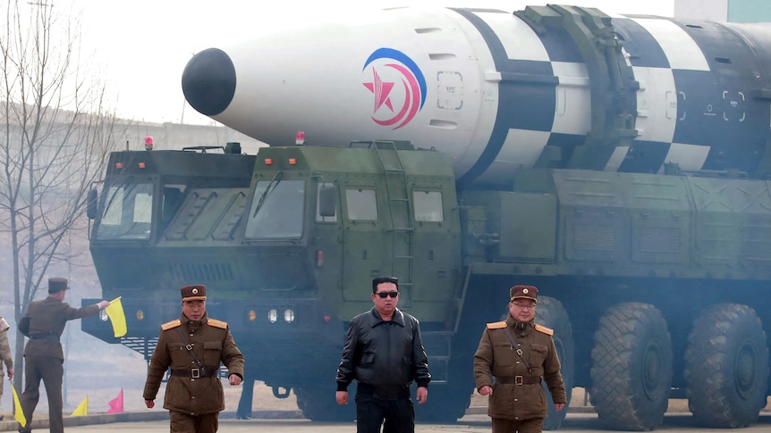 Kim Jong Un and two men in military uniforms walk in front of a military vehicle which is holding a large missile.