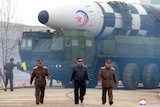 Kim Jong Un and two men in military uniforms walk in front of a military vehicle which is holding a large missile.