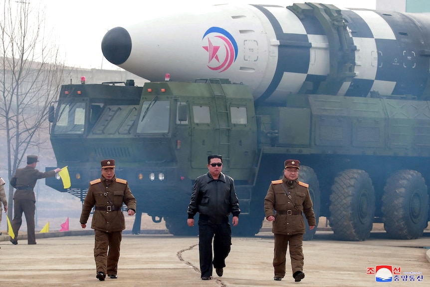 Kim Jong Un e due uomini in uniforme militare camminano davanti a un veicolo militare che trasporta un grosso missile.