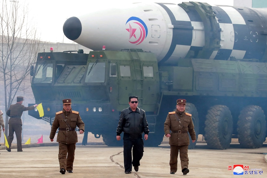 Kim Jong Un and two men in military uniforms walk in front of a military vehicle which is holding a large missile.