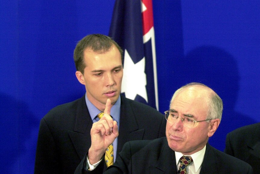 Dutton, still with hair, looks at Howard as he raises a finger, the pair standing in front of an Australian flag.