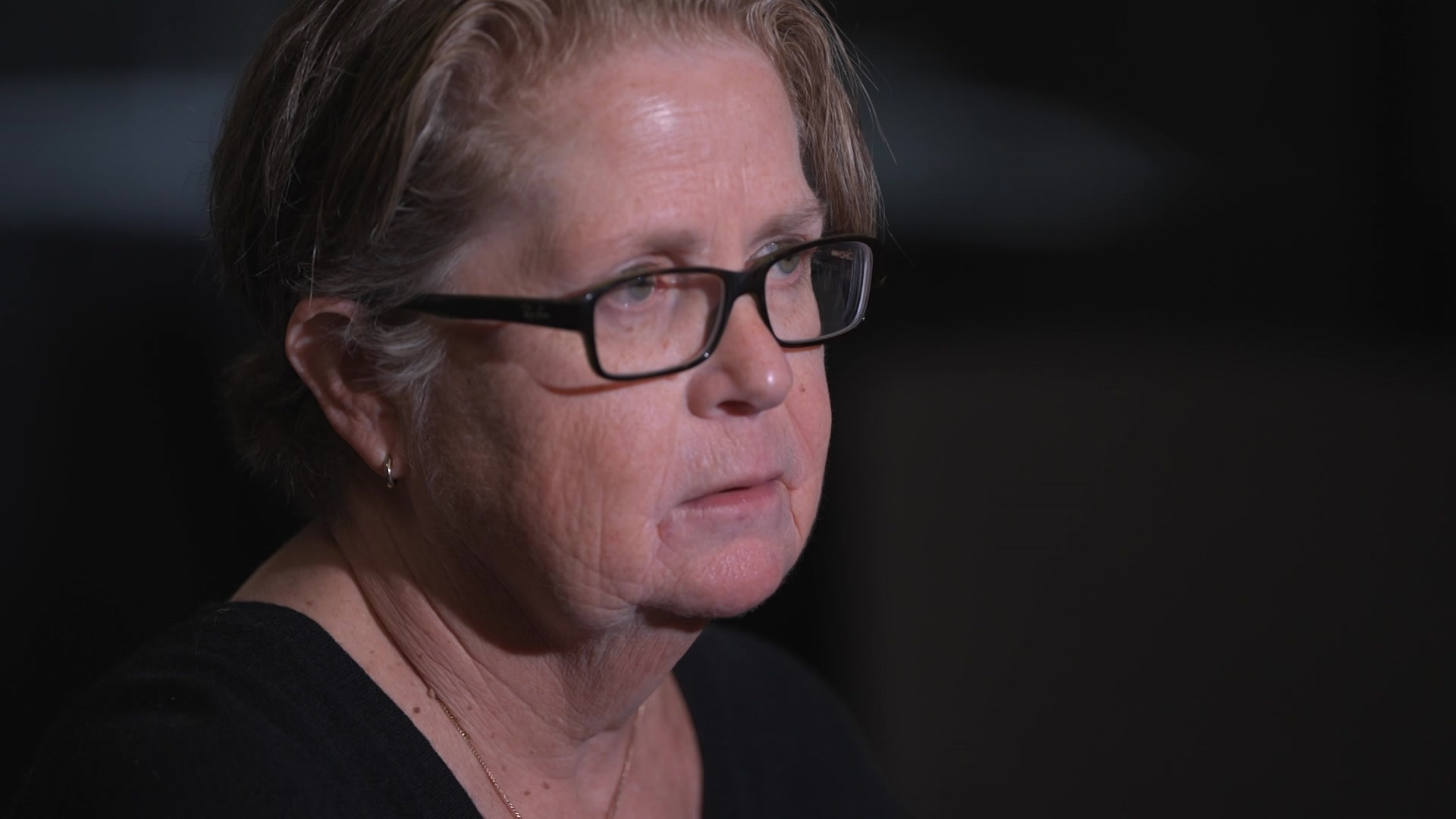 A close-up view of a woman with short blonde hair and black-framed glasses sits in a dark room looking serious.