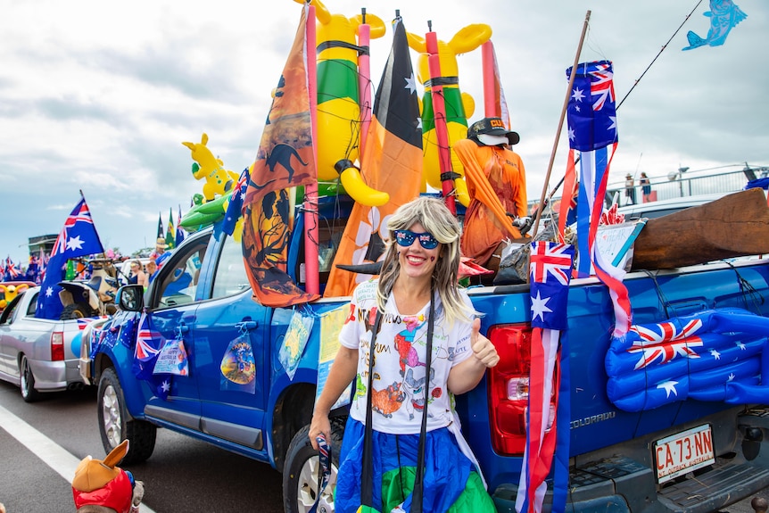 A brightly dressed woman standing in front of a blue ute decorated with Australian flags and mascots, on a track.