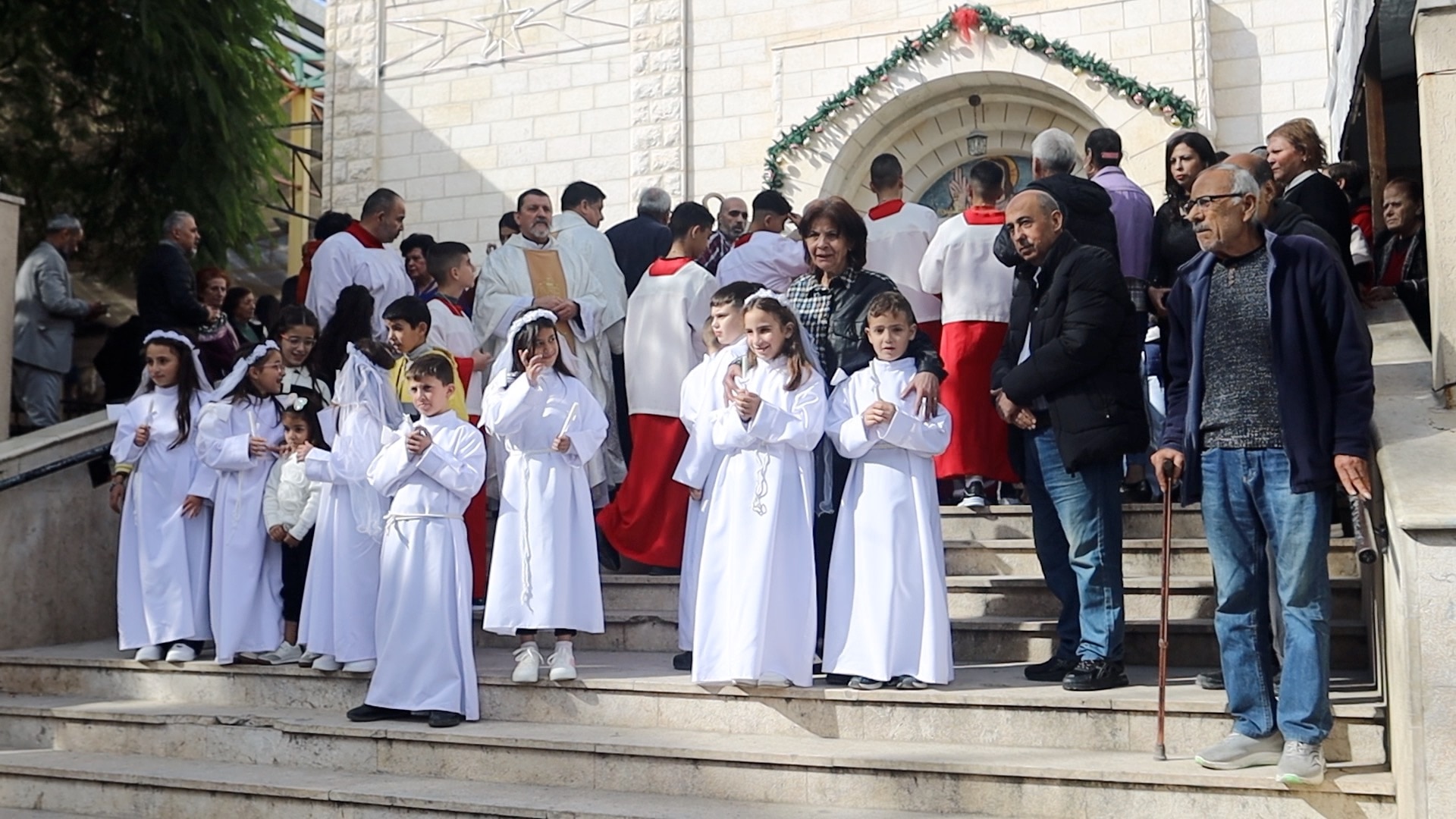 Niños vestidos todos de blanco se encuentran en las escaleras de la iglesia, rodeados de otras personas.