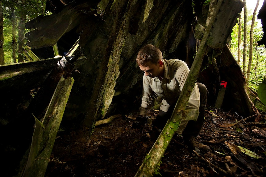 A young man in khaki crouches between the moss covered wreckage of the aircraft 