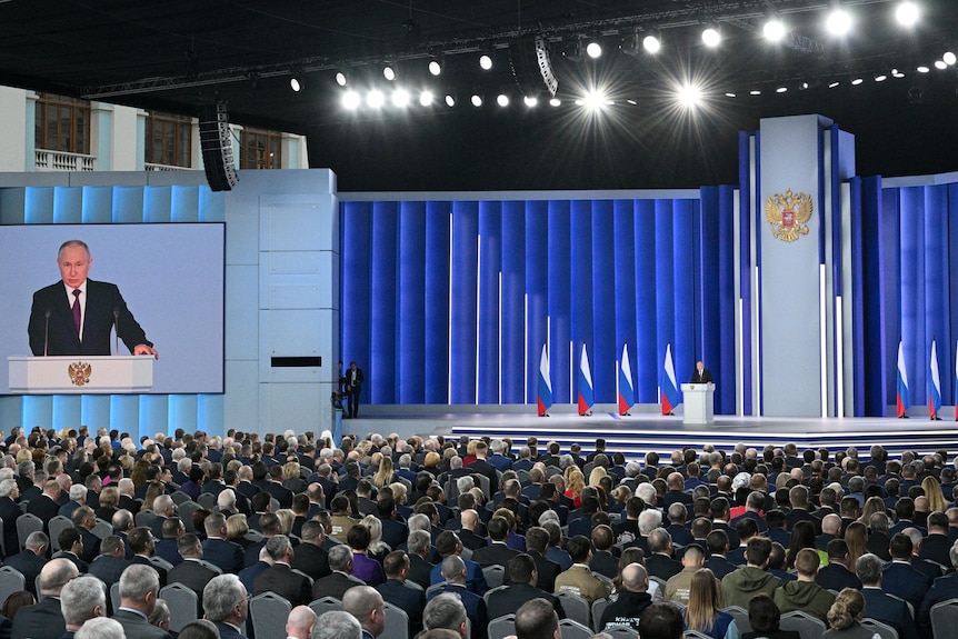 An enormous stage lined with Russian flags. Putin speaks from a lectern and the image is projected on a screen to the left