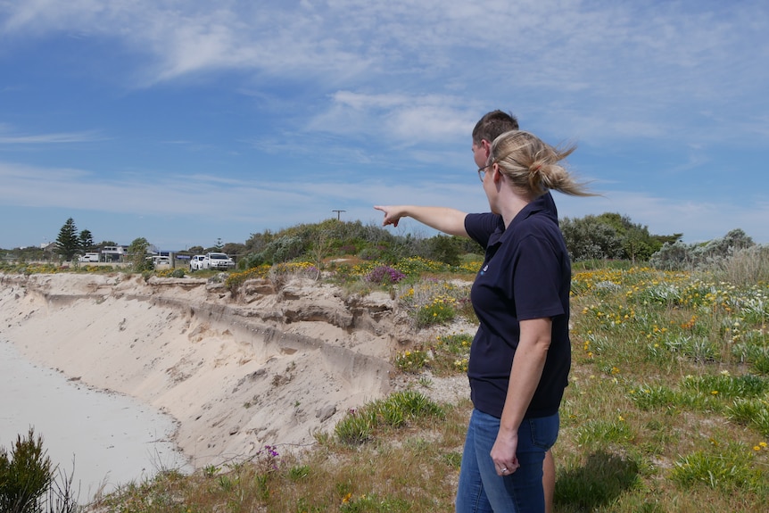 A man and woman standing on a hill, pointing at the beach below. 