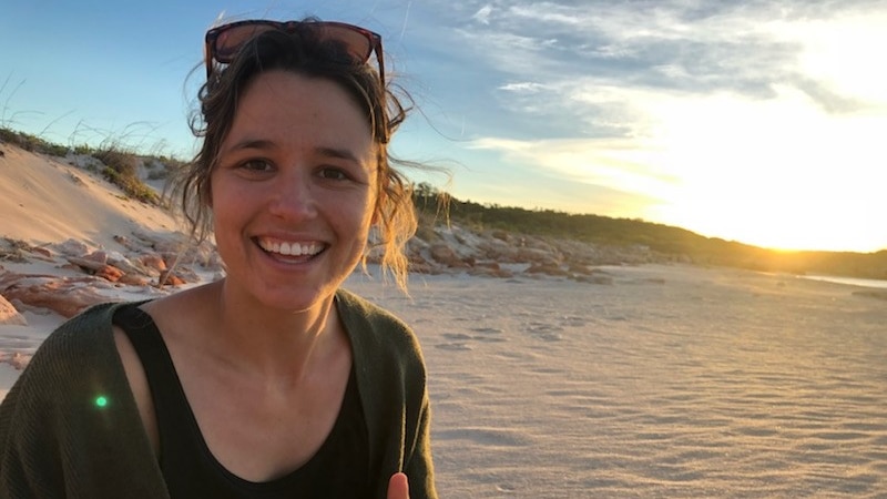 Woman with dark hair smiles while sitting on a beach, with sand and a sunset in the background