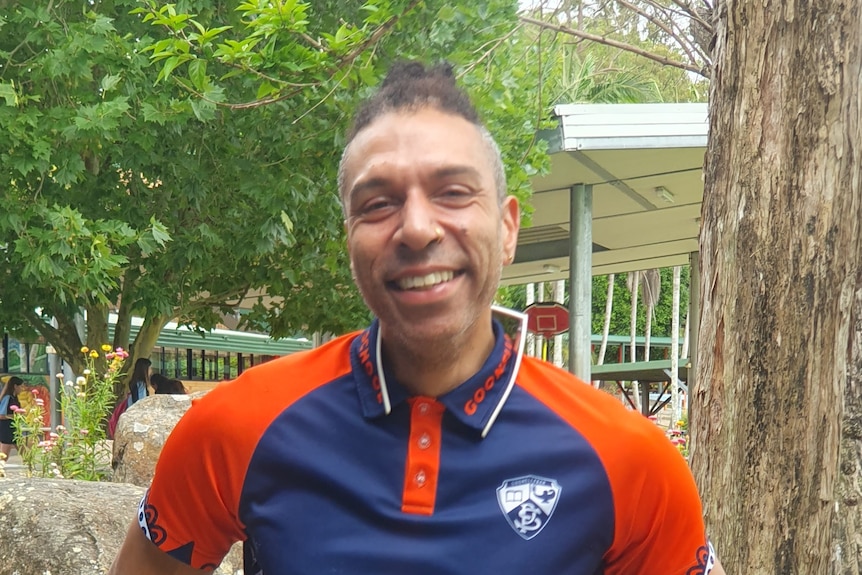 man in his mid thirties wearing a blue and orange shirt, outdoors, by a tree.