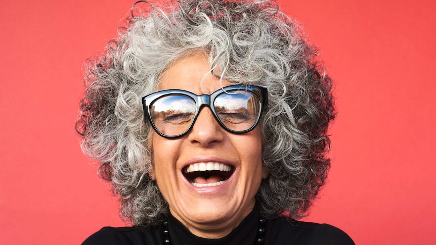 A woman with curly grey hair smiles against a red backdrop.