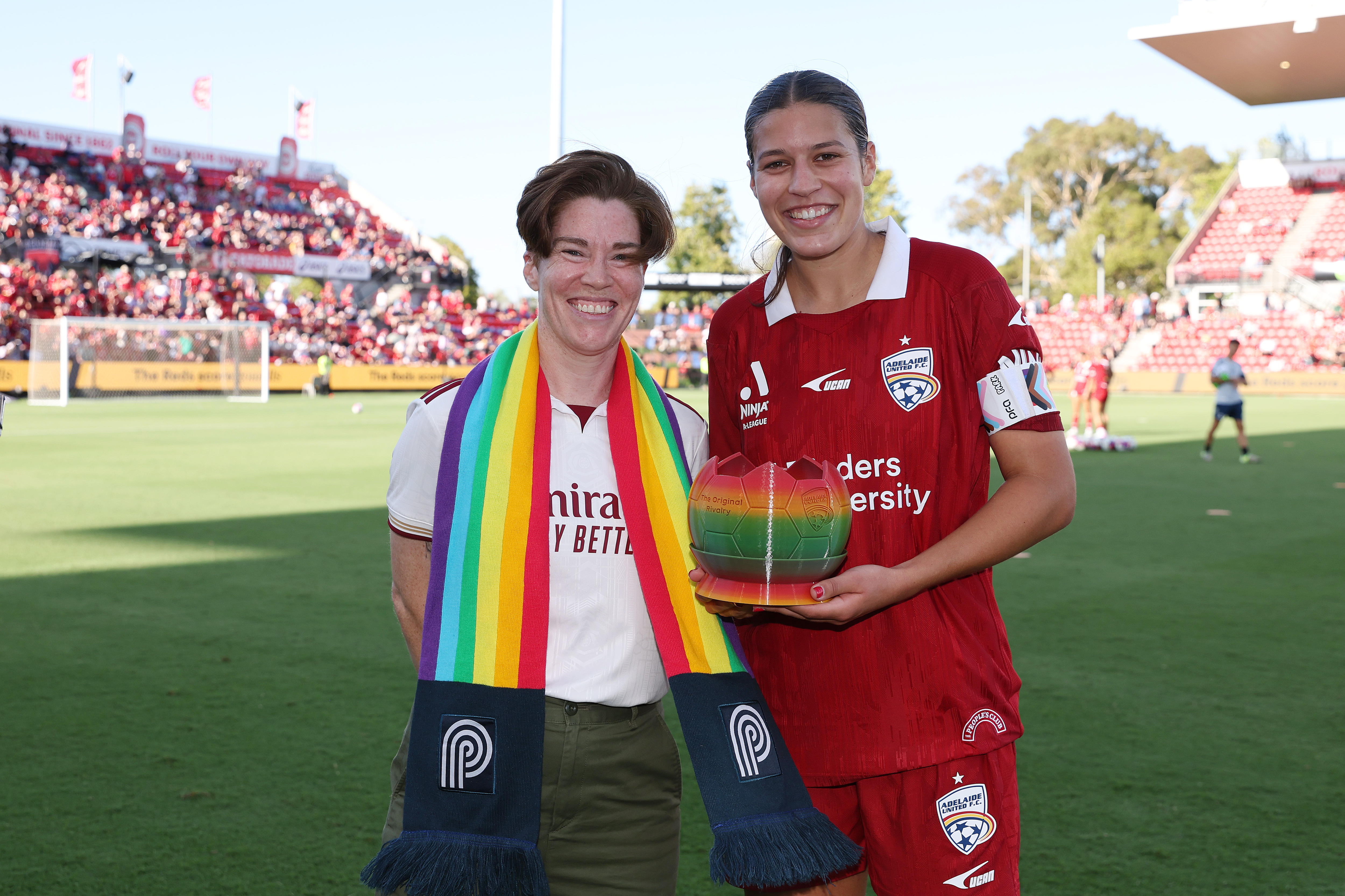 A CEO da Pride Cup, Hayley Conway, usa um lenço arco-íris e fica ao lado da capitã de Adelaide, Ella Tonkin, que segura o troféu da Pride Cup