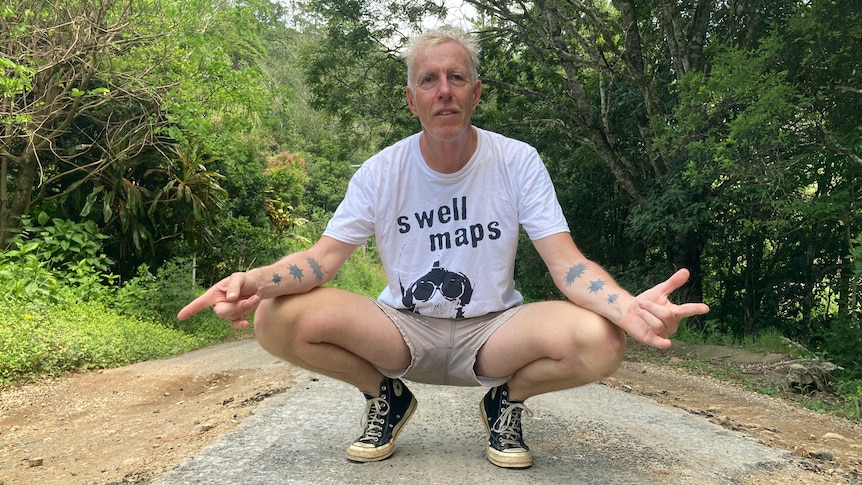 man with white hair and tattoos on his arm  crouched down and pointing to damaged road 