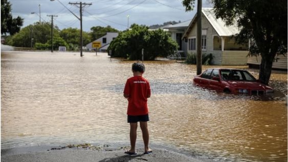 Boy in red tshirt stands on the road in front of flooded street, a car is submerged under water