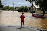 Boy in red tshirt stands on the road in front of flooded street, a car is submerged under water
