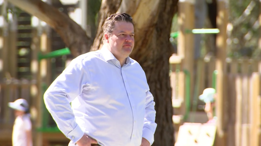 Will Fowles wears a white button up collared shirt and stands with his hands on his hips in front of a tree, looking seriously.