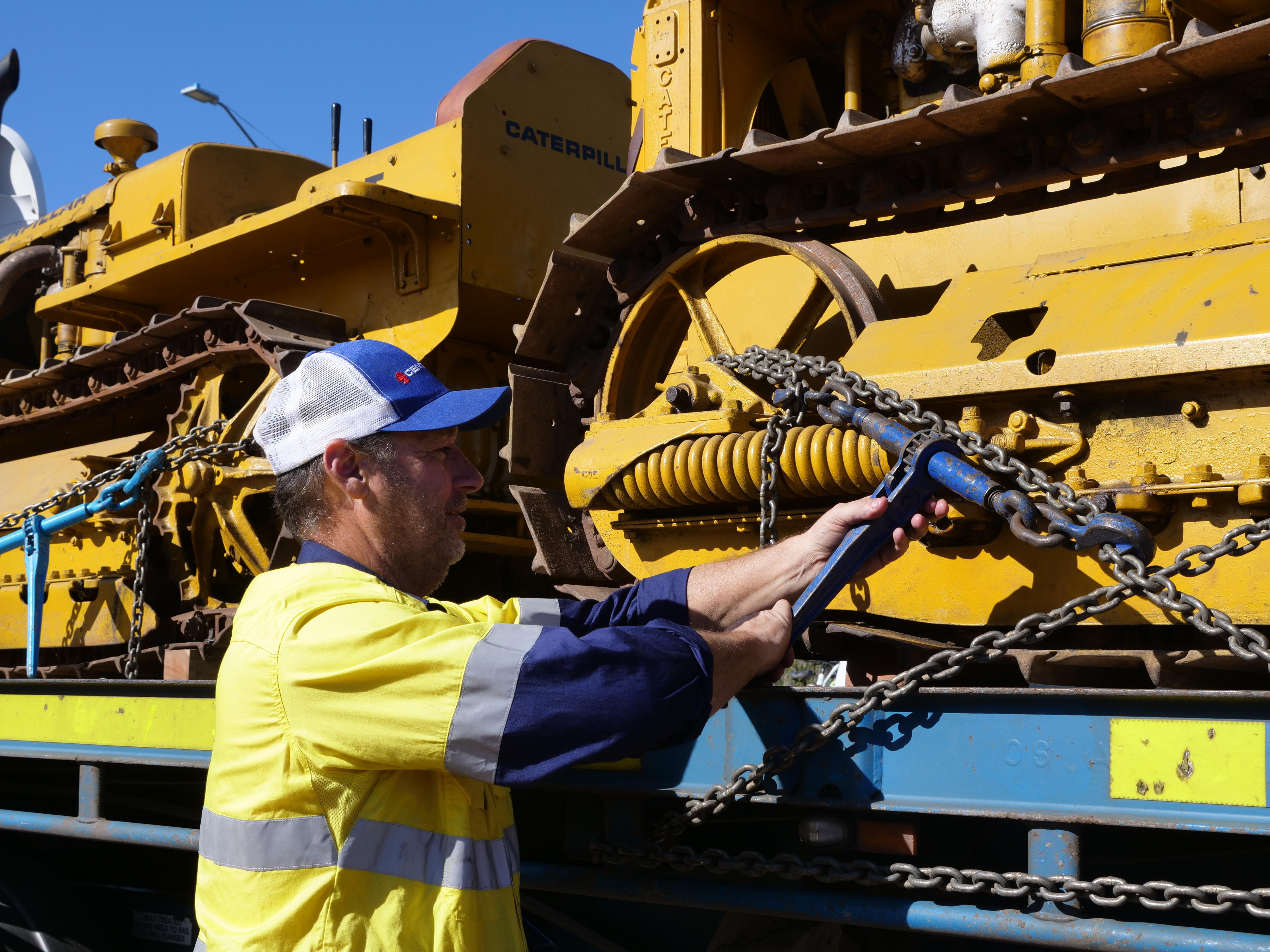 Truck driver adjusts chain holding down yellow bulldozer to truck bed