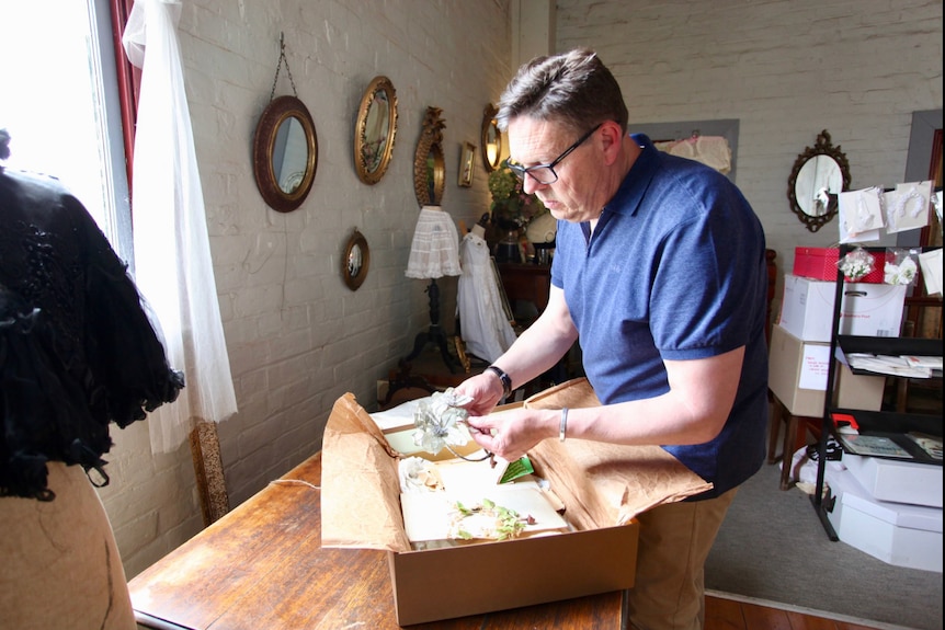 A middle-aged man in a purple t-shirt and glasses examines the contents of a brown cardboard box in a room with vintage mirrors.