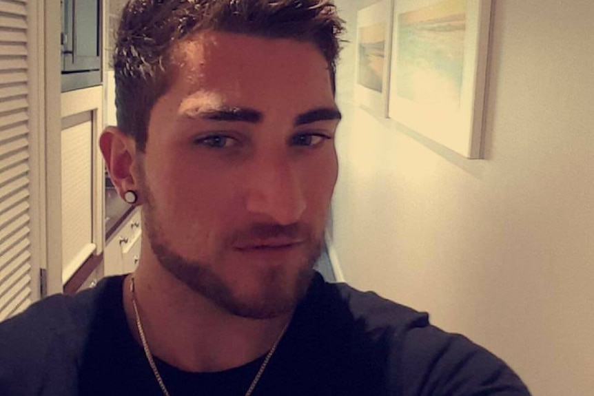 Matthew Fisher-Turner takes a selfie while standing in the hallway of a house wearing a black shirt and neck chain.