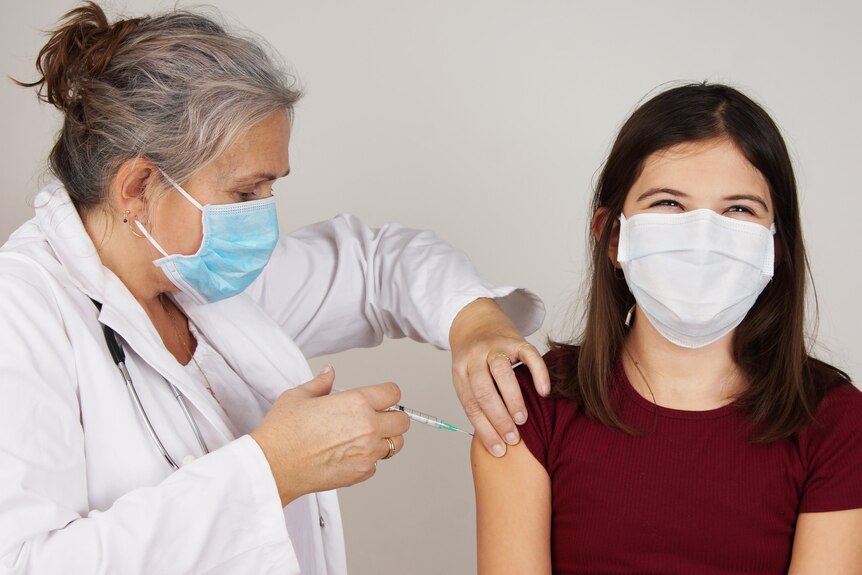 A woman in a white coat and blue face mask vaccinates a teenage girl, who appear to be smiling under her mask.