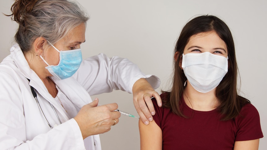 A woman in a white coat and blue face mask vaccinates a teenage girl, who appear to be smiling under her mask.