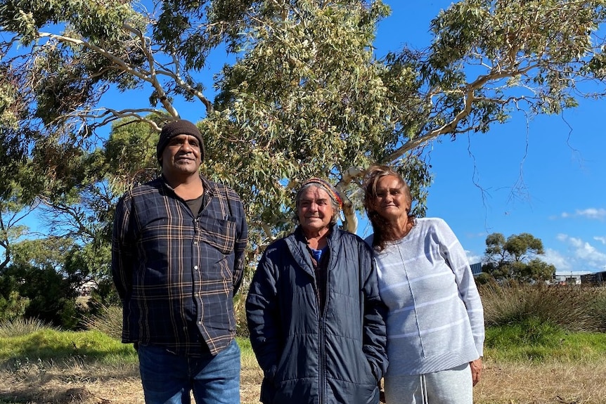 Three First Nation's people standing in front of large gum tree, woman on left two with two men. 