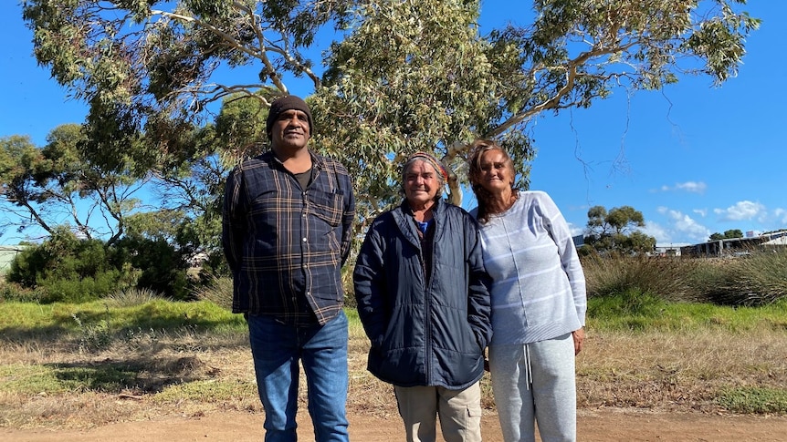 Three First Nation's people standing in front of large gum tree, woman on left two with two men. 