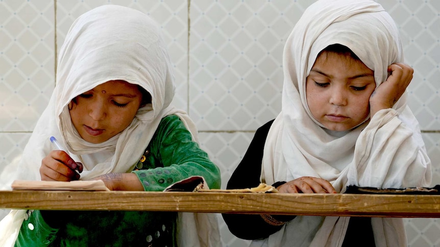 Two young Afghan girls sit at a wooden desk, one writing with a pen and one reading.