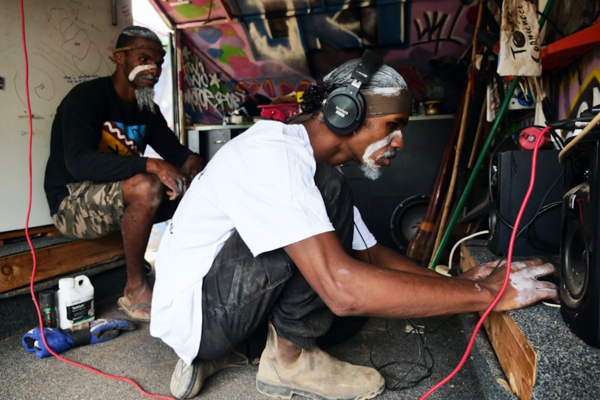 a man in a white shirt with headphones on types on his laptop while another man watches on. they're in a colourful van 