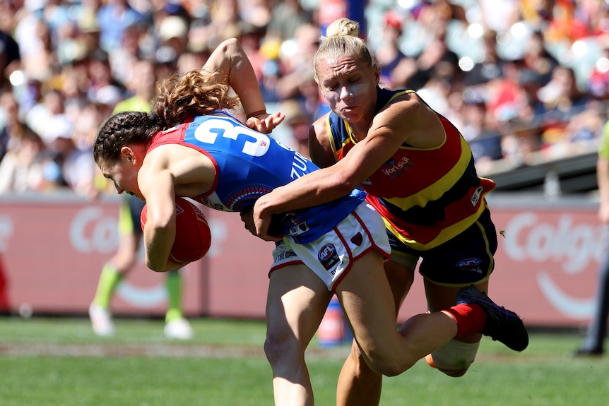 An AFLW player struggles to get free as she carries the ball, while an opponent grabs her in a tackle.