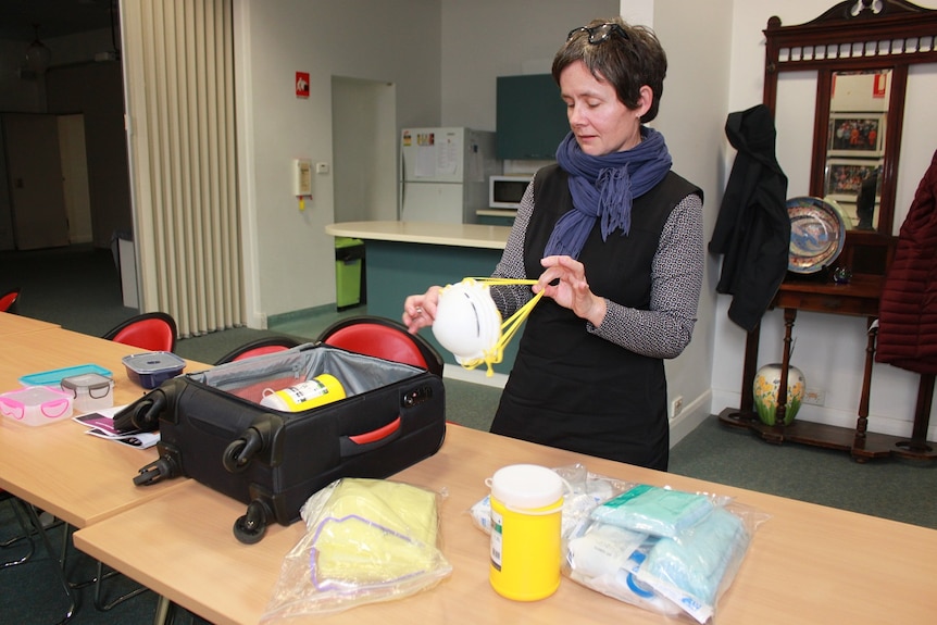 A woman holding a surgical mask stands behind a table with a roller suitcase and items of personal protective equipment.