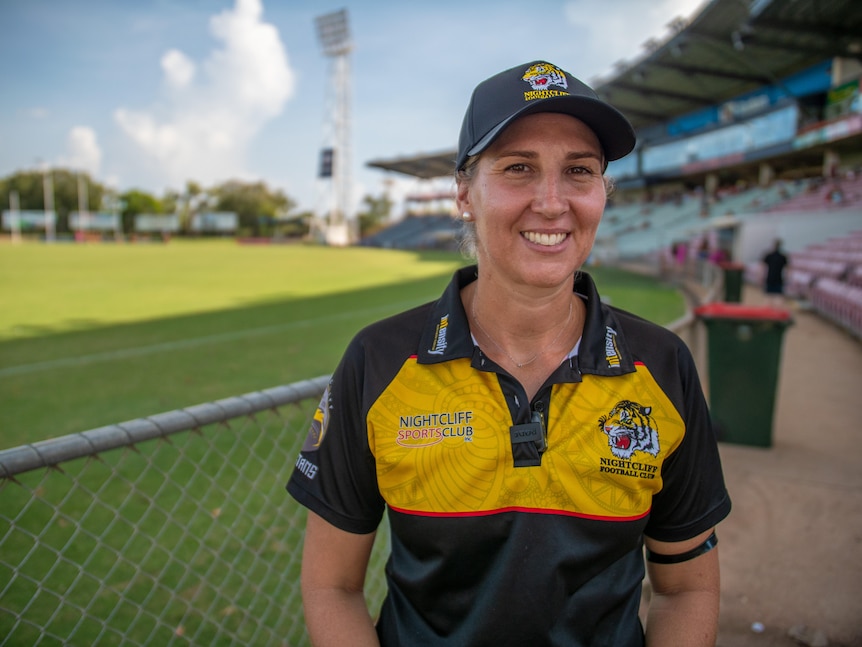 A woman in a team shirt smiles in front of a field and grandstand