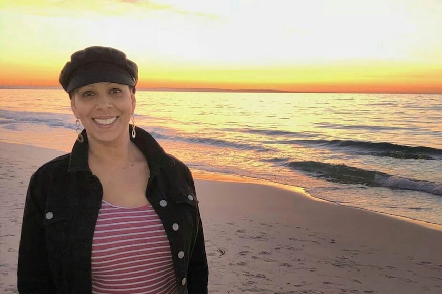 A smiling woman wearing a black cap standing on a beach as the sun sets.