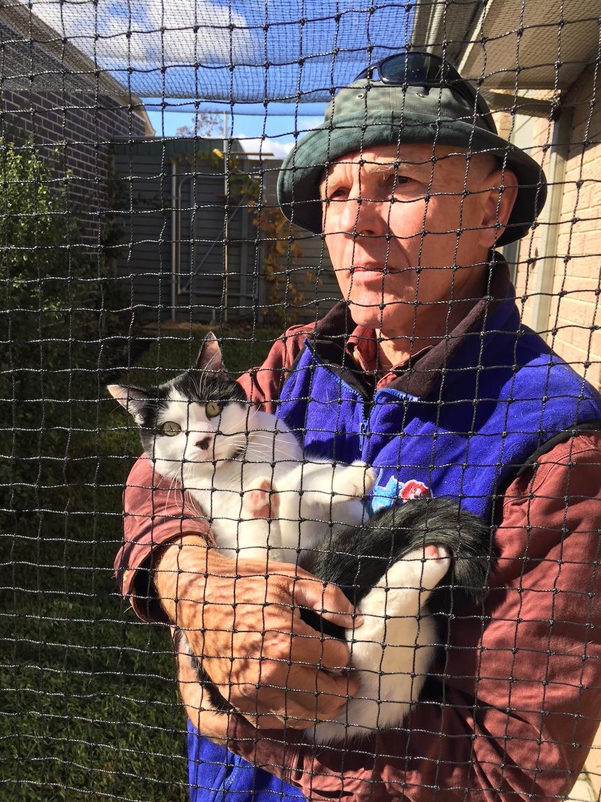 Tom Graham holds a cat behind while standing behind wire mesh.