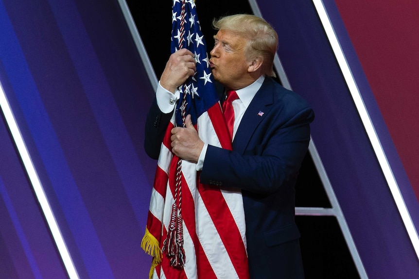 Donald Trump kisses a US flag on a pole, his arms wrapped around the pole in a loving embrace.