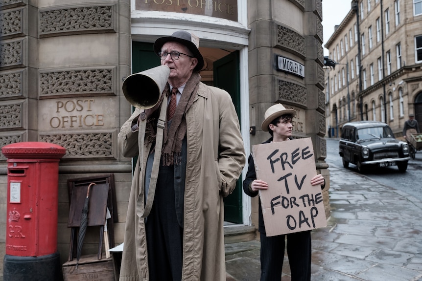 Old man wears taupe trenchcoat and black hat holding megaphone on grey London street beside boy with 'free TV for the OAP' sign