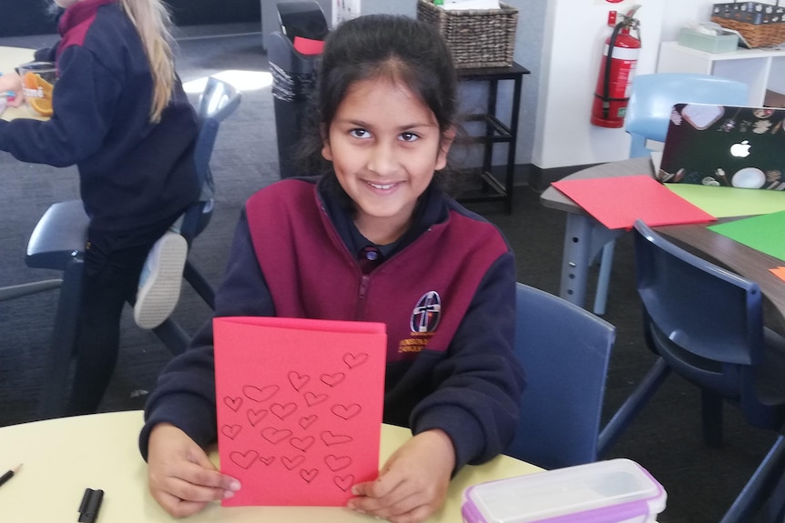 A girl sitting at a desk holds up a handmade card with pictures of love hearts on the front