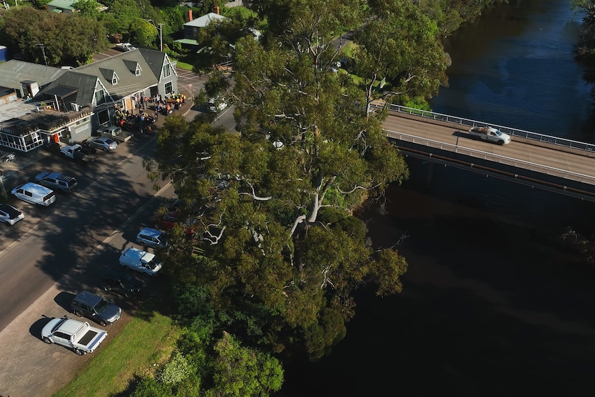 Drinkers outside a country pub viewed from the air, a ute crossing a river bridge nearby