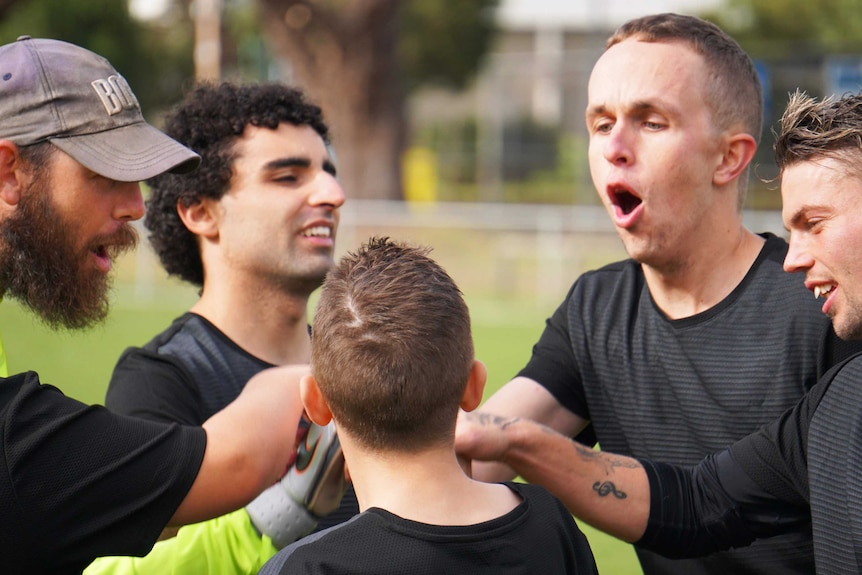 Four men and one boy in black soccer jerseys pile hands in a circle during a pre-game chant on the pitch.