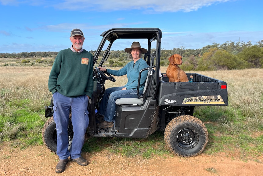 A woman sits behind the steering wheel of a small electric farm vehicle with a dog in the back. A man stands beside the vehicle.