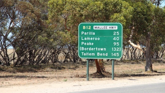 A green sign with town names is on the side of a road. There are trees behind it.