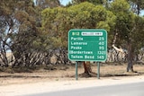 A green sign with town names is on the side of a road. There are trees behind it.