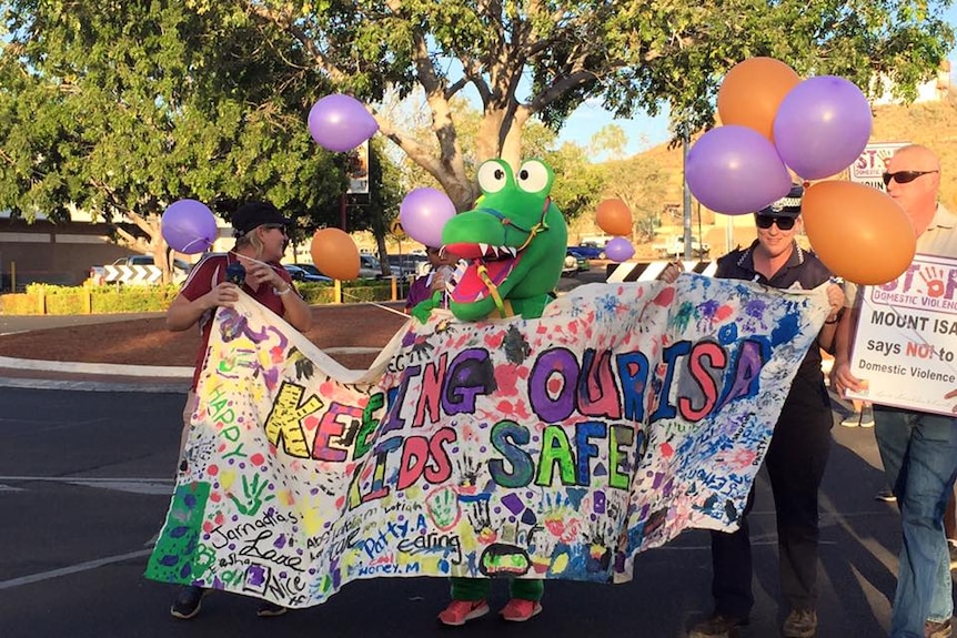 A police woman and a crocodile mascot hold a colourful banner reading 'keeping our kids safe'.
