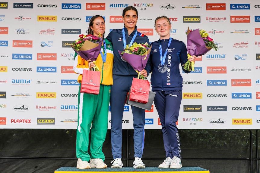 Three kayak cross athletes stand on a podium wearing medals around their necks, holding flowers, and smiling.