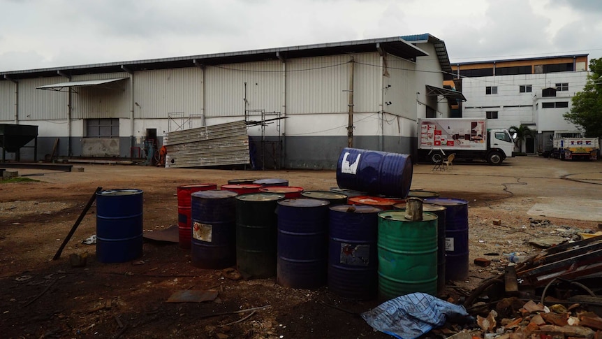 A picture of red, blue and green barrels outside a corrugated iron warehouse on a dirt ground, trucks in the background.