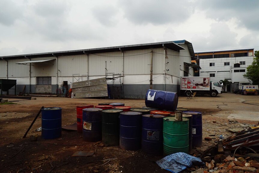 A picture of red, blue and green barrels outside a corrugated iron warehouse on a dirt ground, trucks in the background.