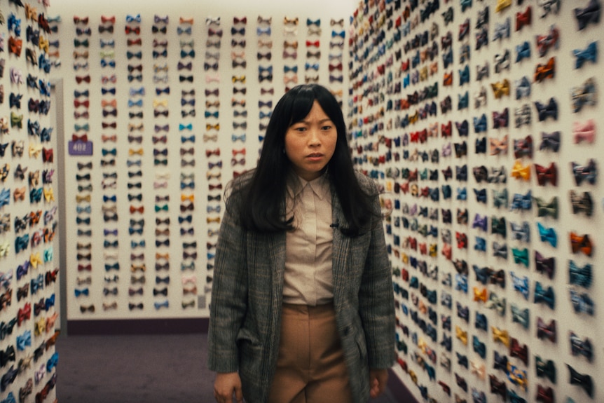 Awkwafina in a long wig looks stressed in a room with the walls lined by bowties.