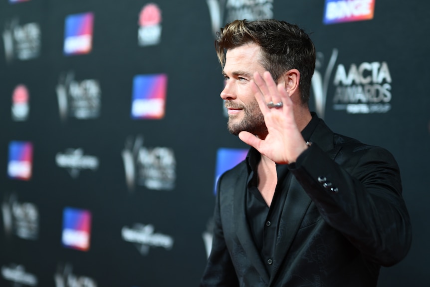 Chris Hemsworth, a white man with blonde hair, wears a sleek black suit and waves to photographers on a red carpet.